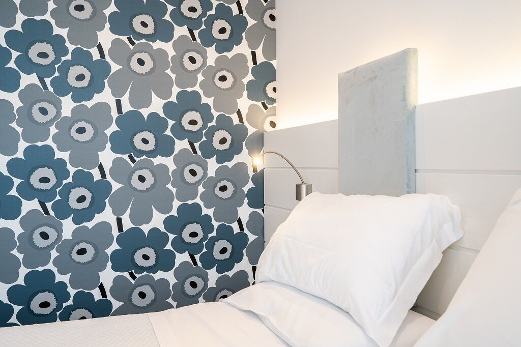 Habitación de hotel con pared de estampado floral, cama con ropa blanca y mesita de noche moderna con lámpara.