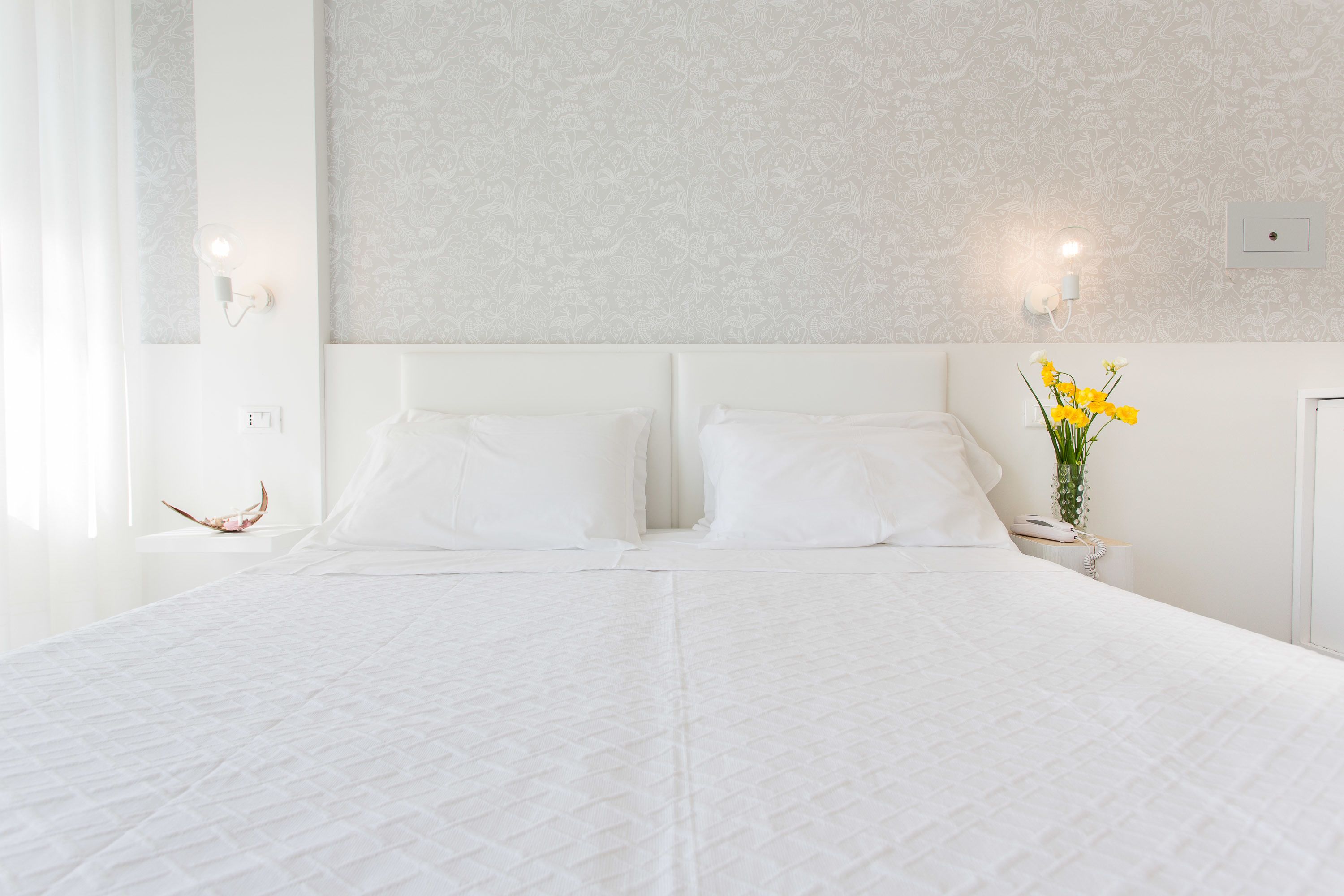 A large bed with white bedding, a nightstand with a vase of yellow flowers, and a wall-mounted light fixture.