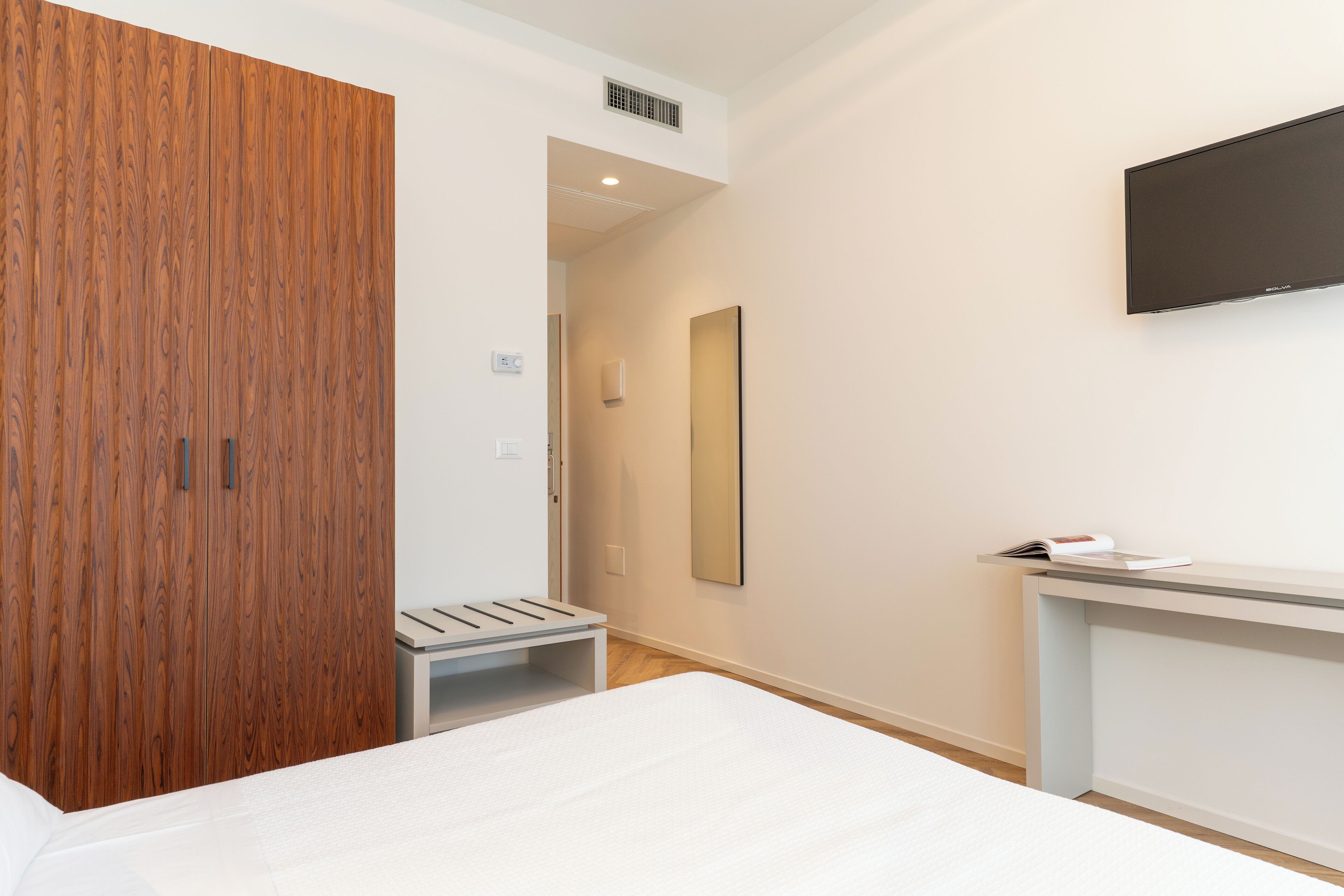 A hotel room with a wooden wardrobe, a bed, a bedside table with a magazine, a mirror, and a TV mounted on the wall.