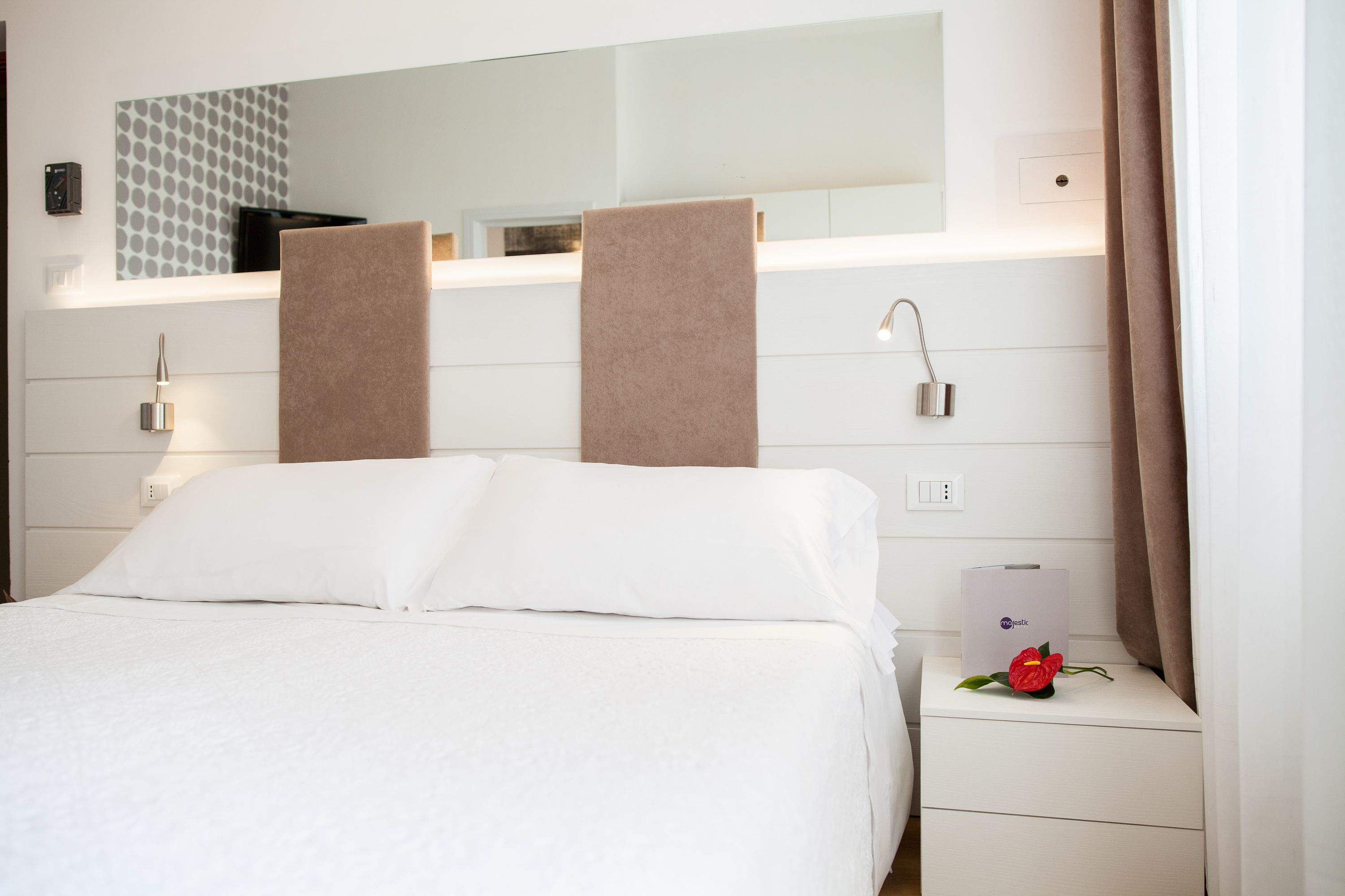 Habitación de hotel con cama, mesita de noche con lámpara, espejo y una flor roja sobre la mesita.