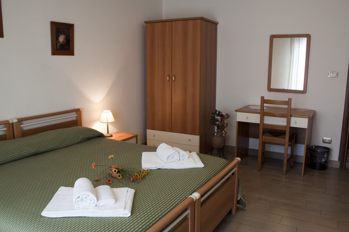 Ein ordentlich bezogenes Bett mit aufgerollten Handtüchern, ein Holzschrank, ein Schreibtisch mit Stuhl und ein Spiegel.