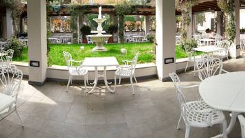 A covered outdoor seating area with white metal tables and chairs, a central fountain, and a well-maintained lawn.
