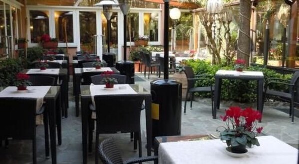 Un área de comedor al aire libre con mesas y sillas, rodeada de plantas y un recinto de cristal.