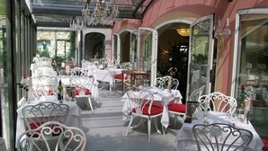 An outdoor dining area with white metal chairs and tables, covered by a glass roof, and a pink building in the background. - Hotel Due Mari (Sestri Levante)