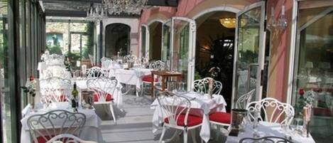 An outdoor dining area with white metal chairs and tables, covered by a glass roof, and a pink building in the background.