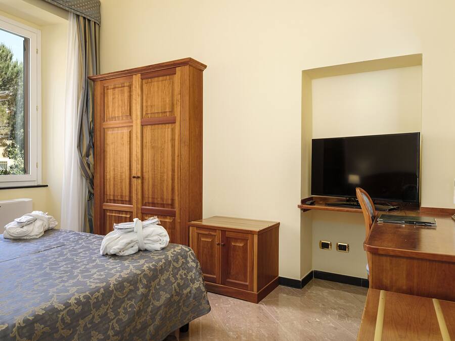 A hotel room with a bed, a wooden wardrobe, a desk with a television, and a window with curtains.
