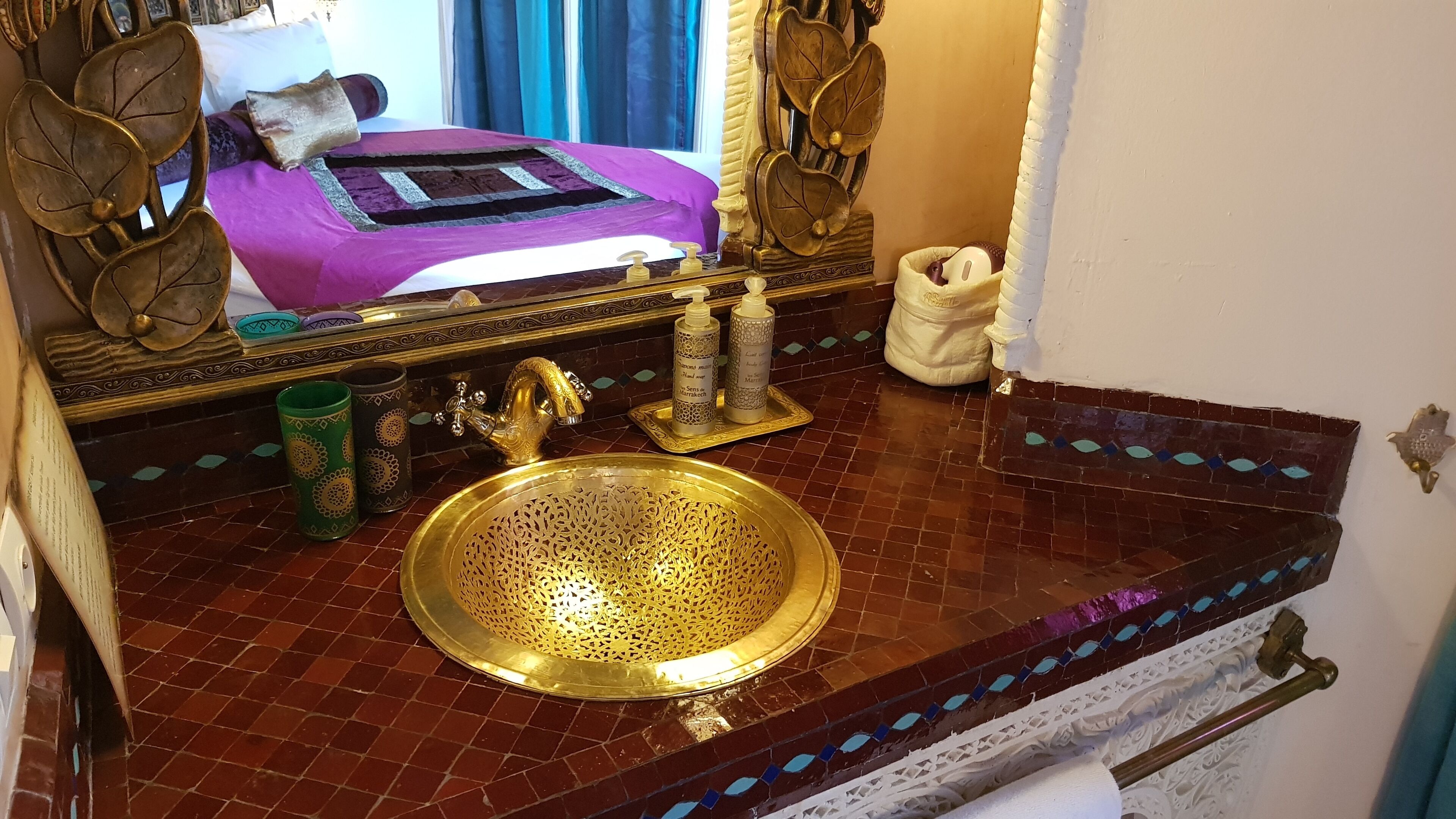 Une salle de bain avec un lavabo doré, un miroir et un lit recouvert d’une couverture violette.