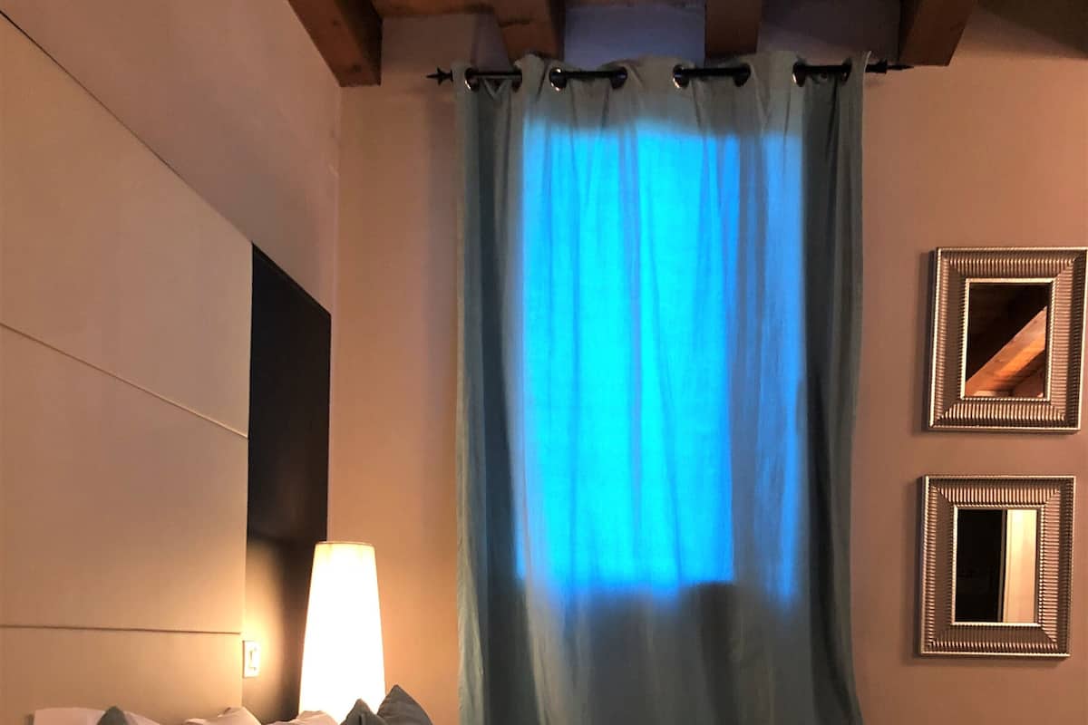 A hotel room with a bed, pillows, a lamp, a mirror, and a window with blue curtains.