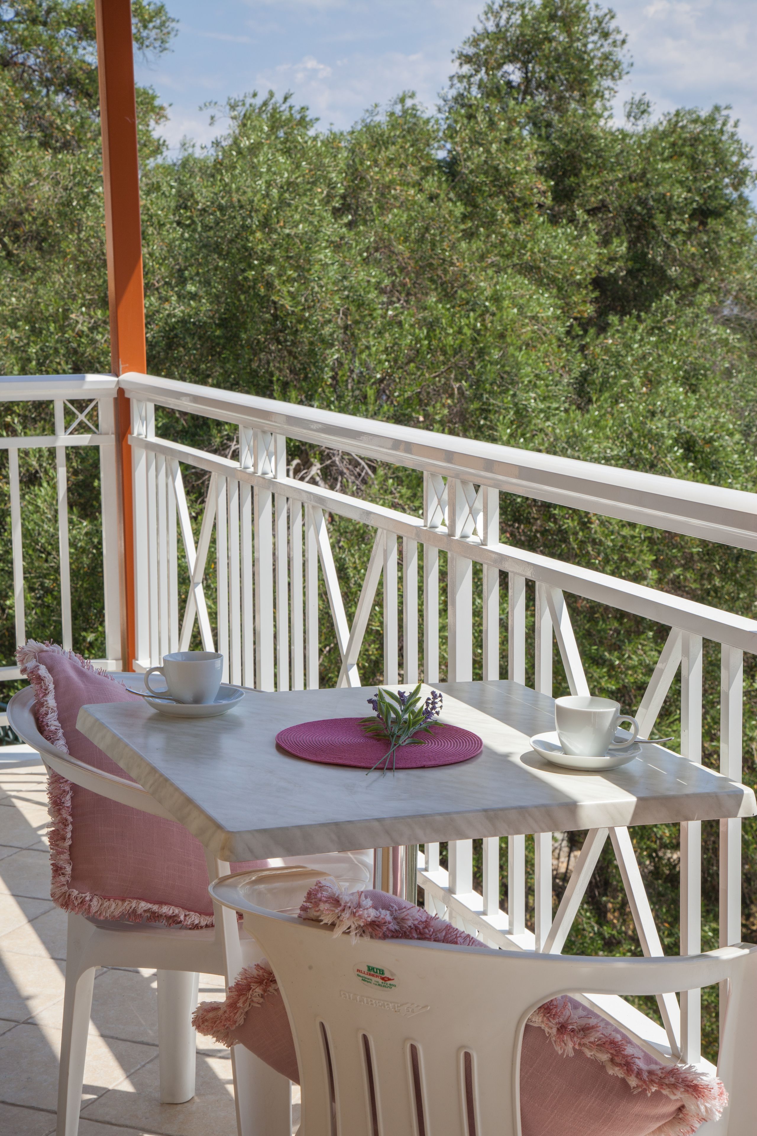 A white outdoor seating area with a table and chairs, surrounded by a white railing and greenery in the background.