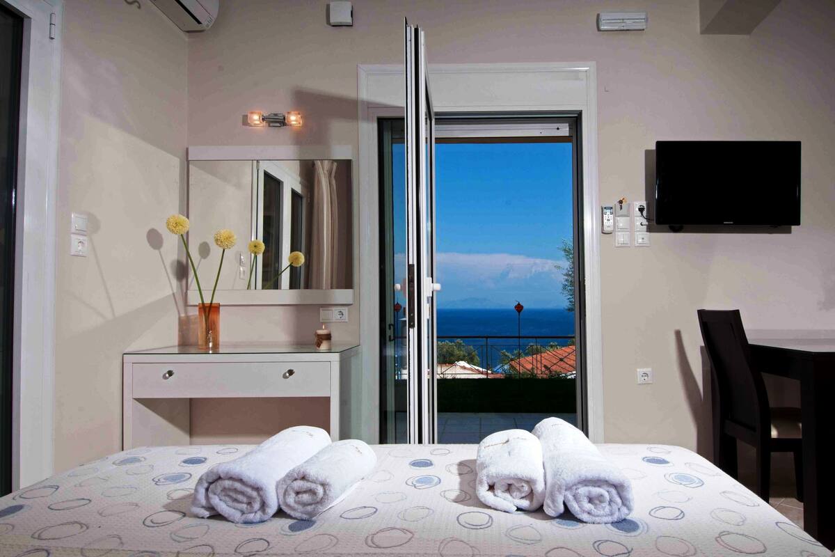A hotel room with a bed, a desk, a mirror, a vase with flowers, and a view of the sea.