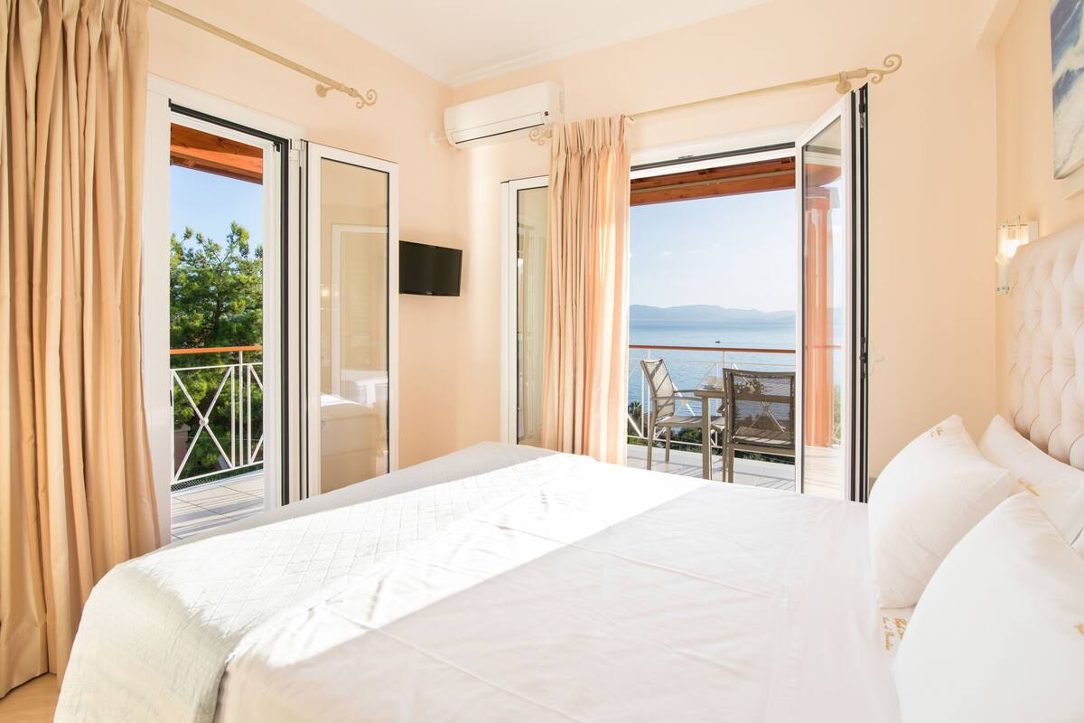 A bedroom with a large bed, a TV, and a balcony with a view of the sea.
