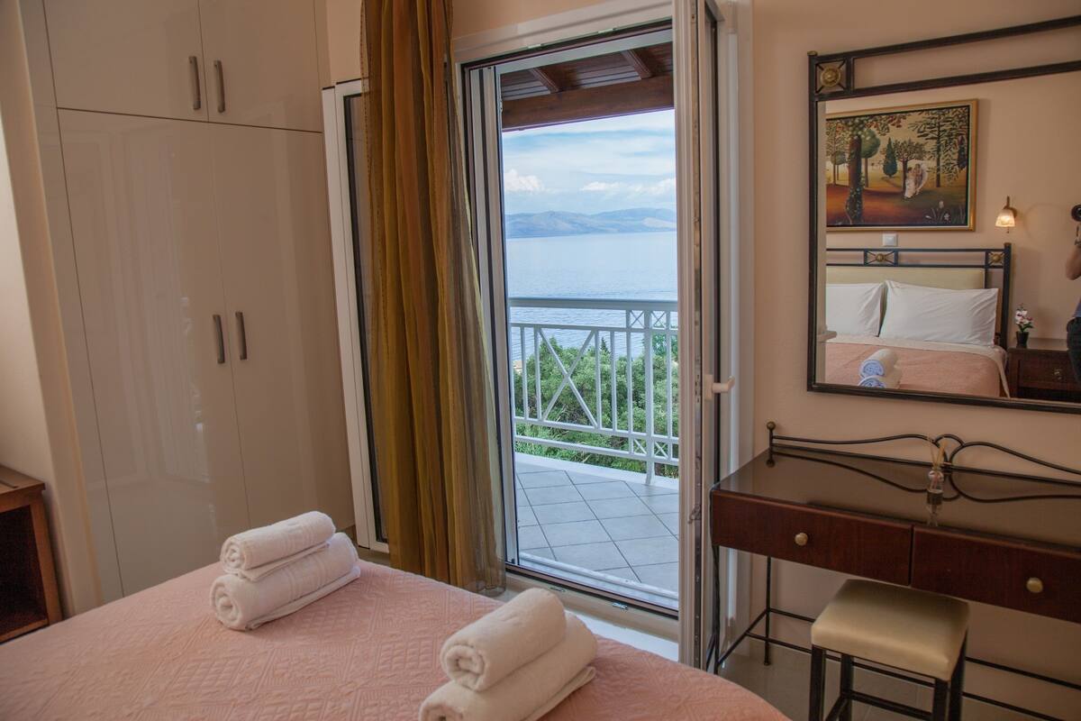 A bedroom with a bed, a desk, and a view of a balcony with a railing.