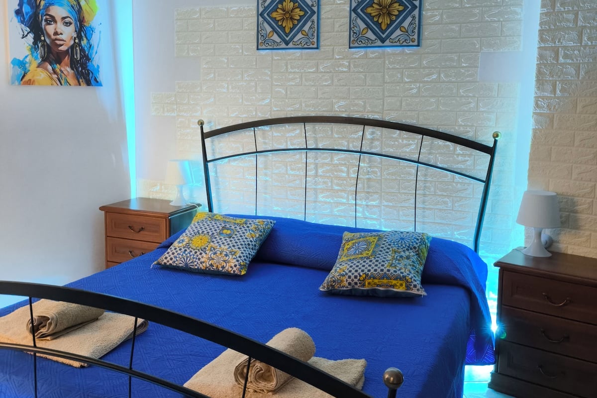 A bedroom with a blue bedspread, two patterned pillows, a blue headboard, a nightstand with a lamp, and a wall with decorative tiles and a painting.