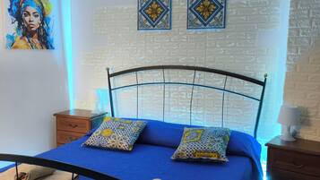 Een slaapkamer met een blauw beddekleed, twee kussens met dessin, een blauw hoofdbord, een nachtkastje met een lamp, en een muur met decoratieve tegels en een schilderij.