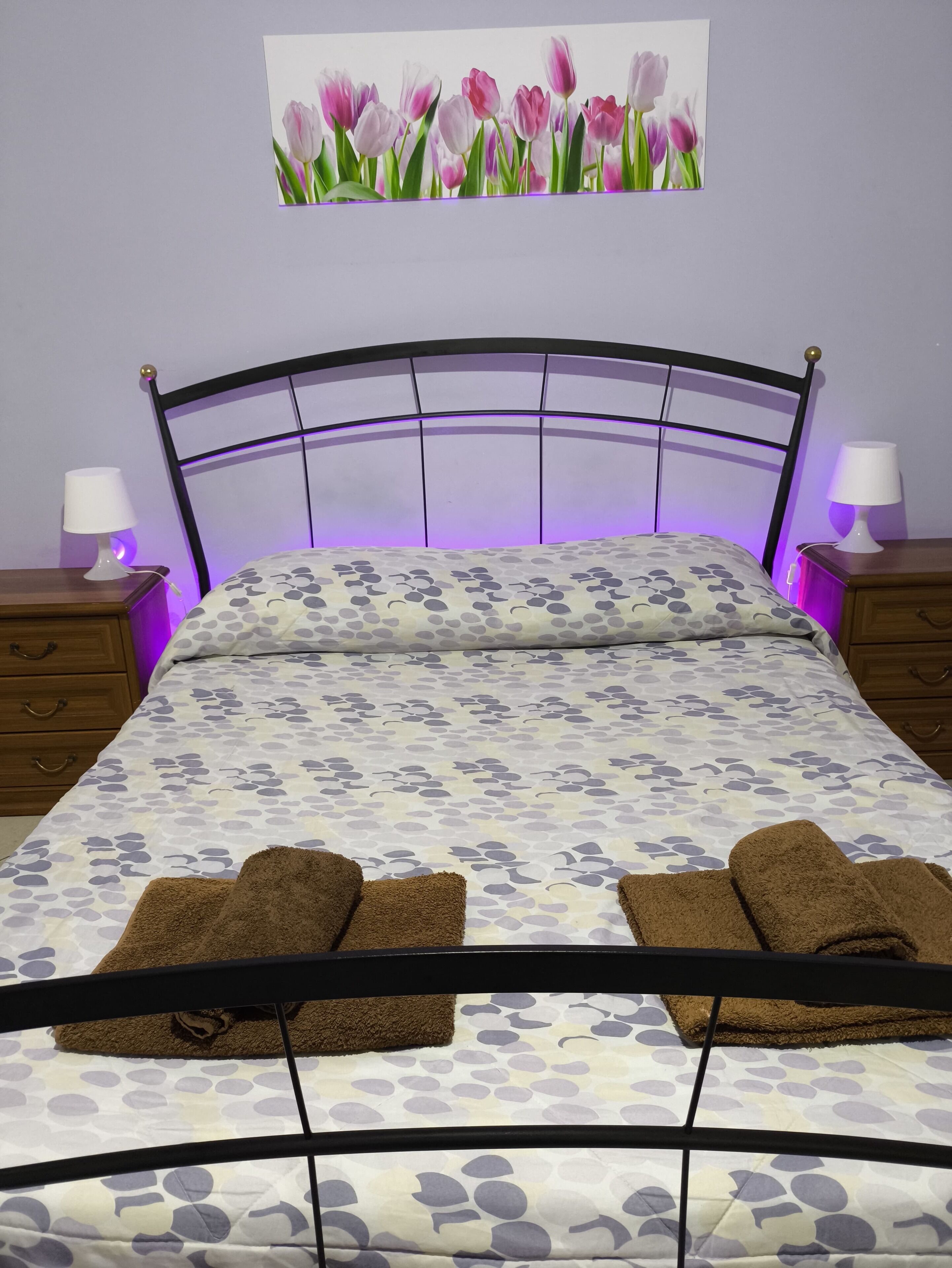 Ein Bett mit einem blumigen Überwurf, zwei braune Handtücher, ein Kopfteil mit violetten Licht und ein Gemälde von Tulpen an der Wand.