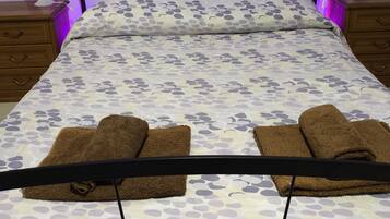 Ein Bett mit einem blumigen Überwurf, zwei braune Handtücher, ein Kopfteil mit violetten Licht und ein Gemälde von Tulpen an der Wand.