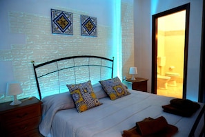 A bedroom with a brick-patterned wall, a bed with patterned pillows, a nightstand with a lamp, and a bathroom visible through an open door.
