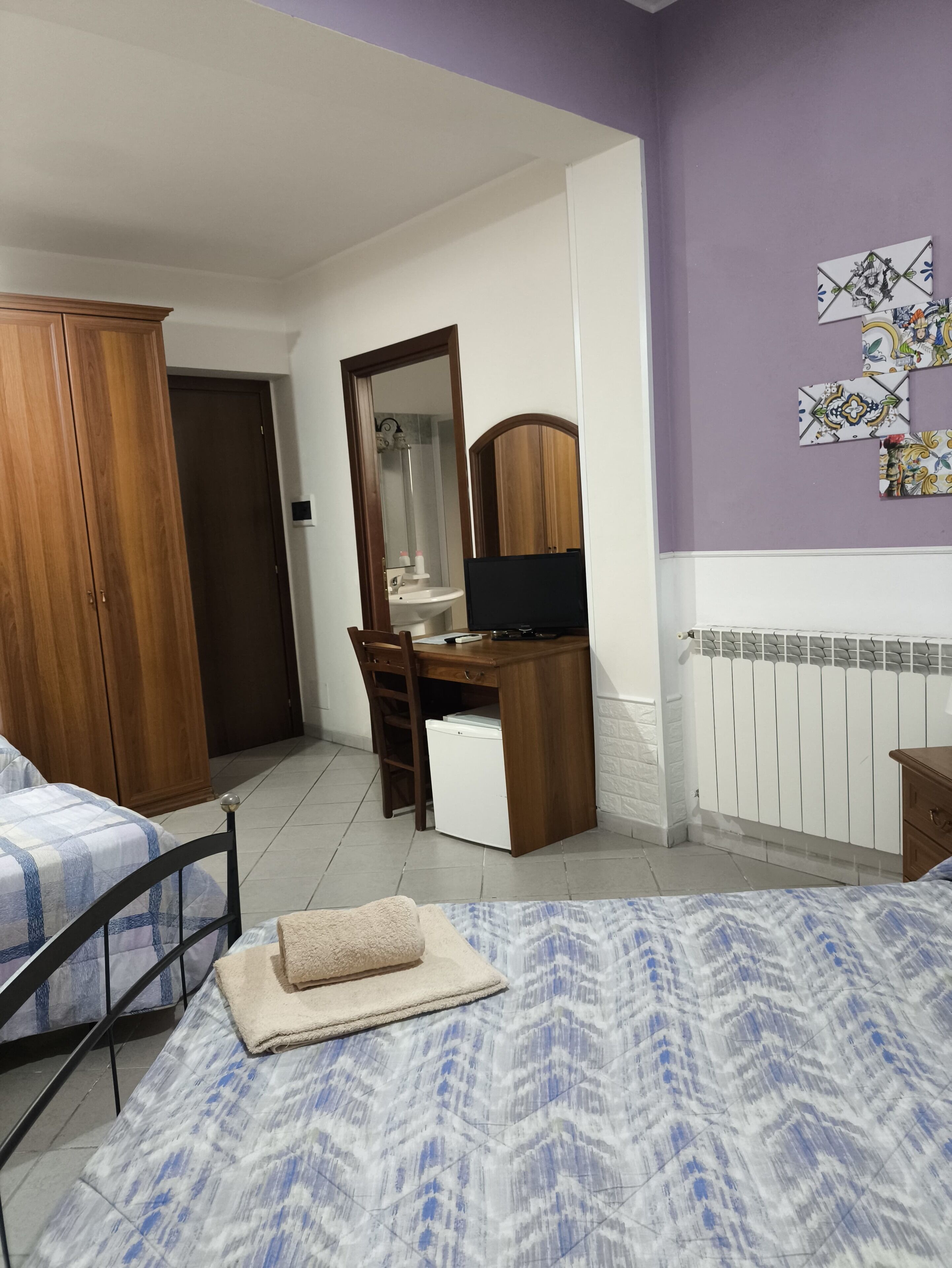 Quarto com cama, guarda-roupa de madeira, escrivaninha com cadeira, espelho e radiador.