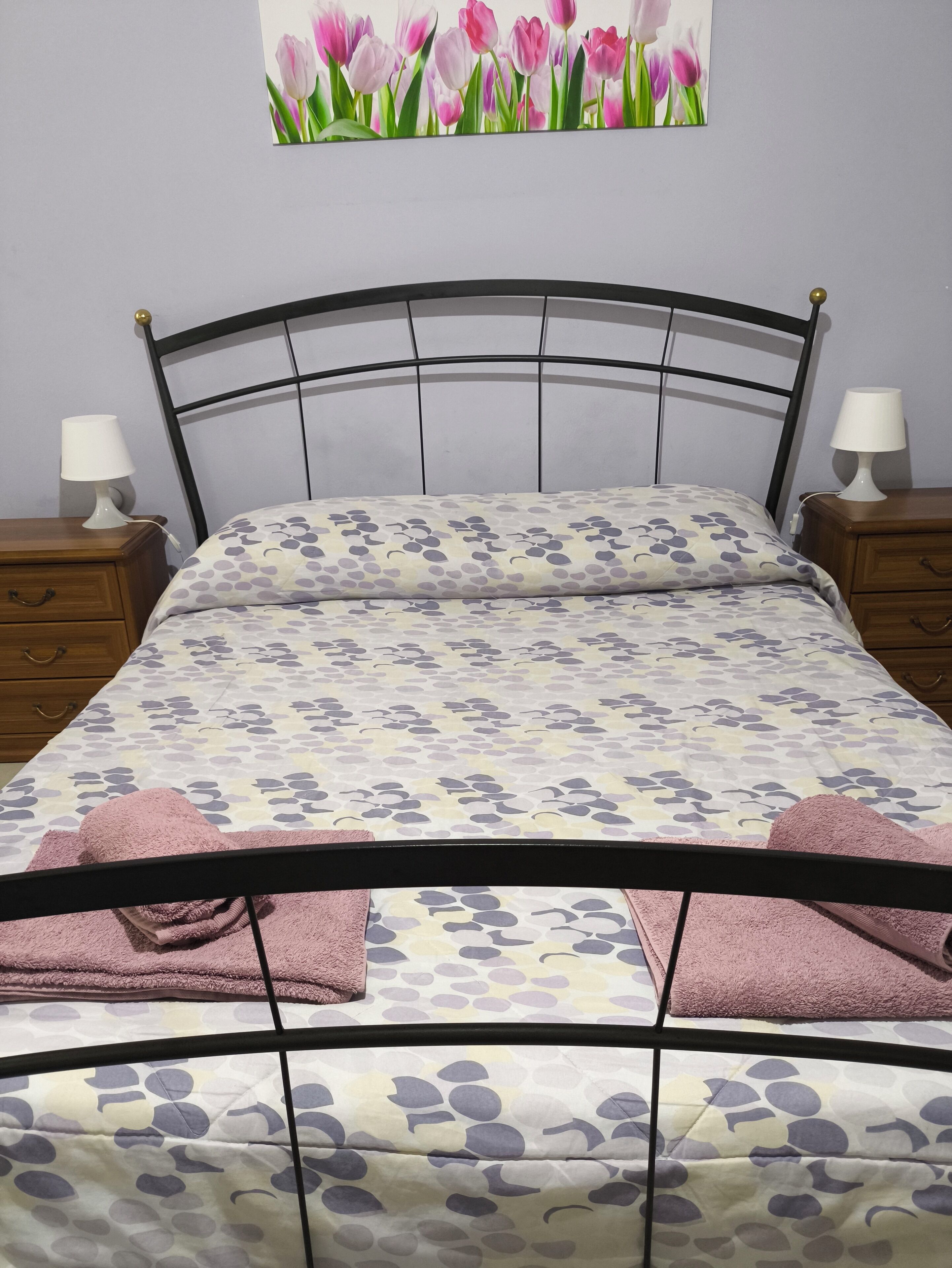 Ein Bett mit floralem Überwurf, zwei Nachttische mit Lampen und ein Wandbild von rosafarbenen Tulpen.