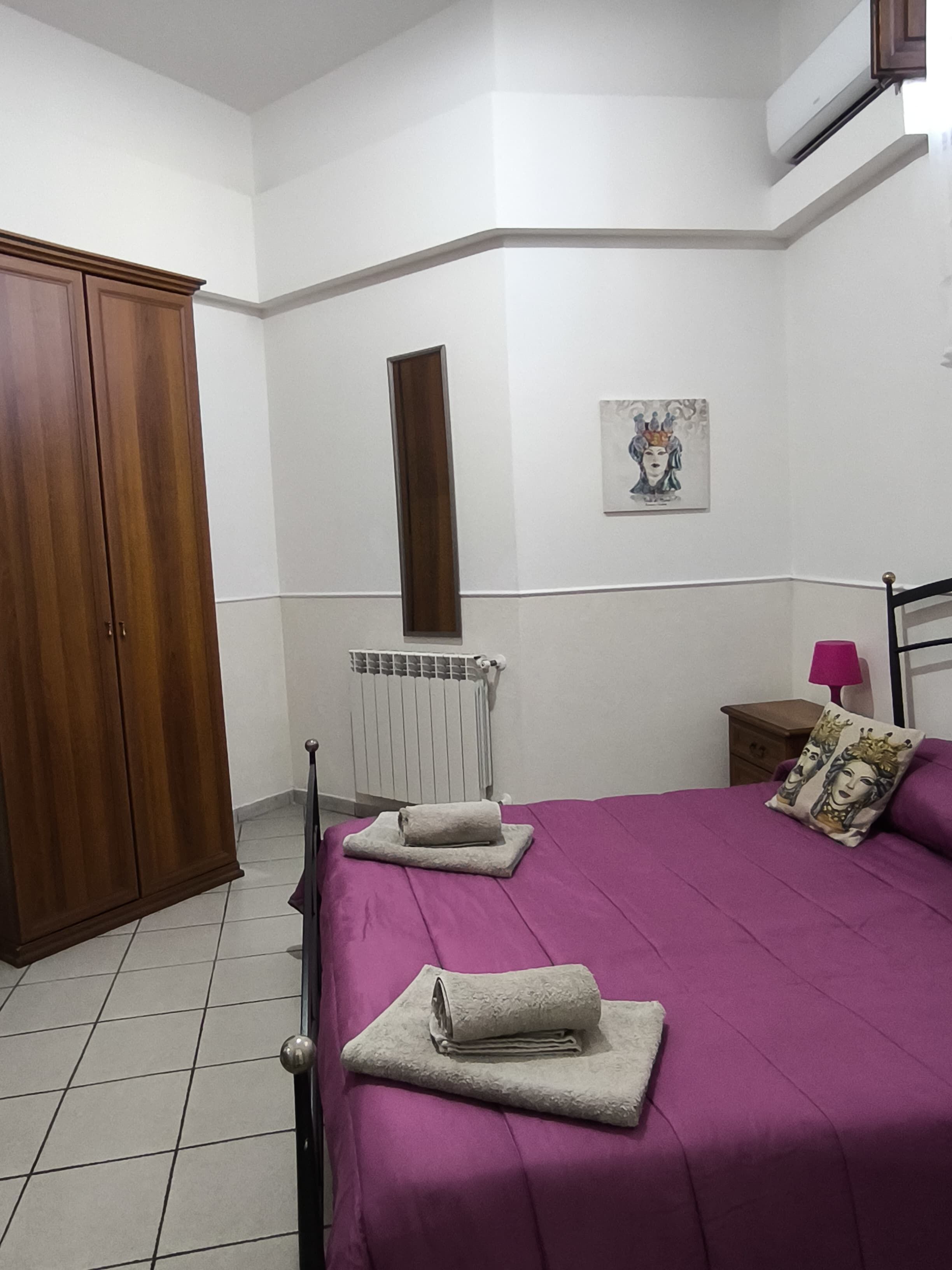 A bedroom with a bed, a wardrobe, a radiator, a nightstand, a lamp, and a painting on the wall.