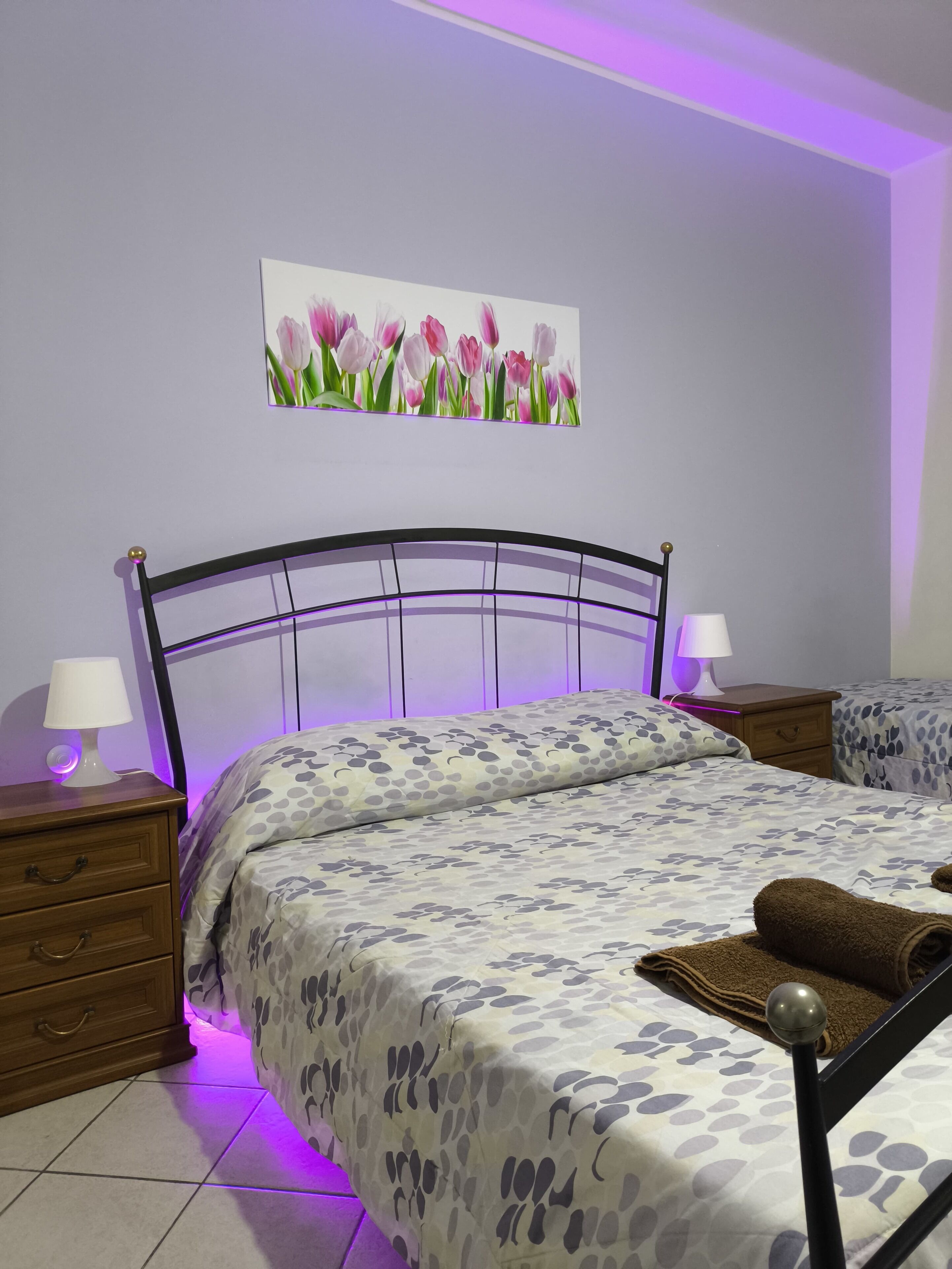 Une chambre à coucher comprenant un lit, deux tables de chevet, une petite table d’appoint, un tableau accroché au mur et un éclairage d’ambiance violet.