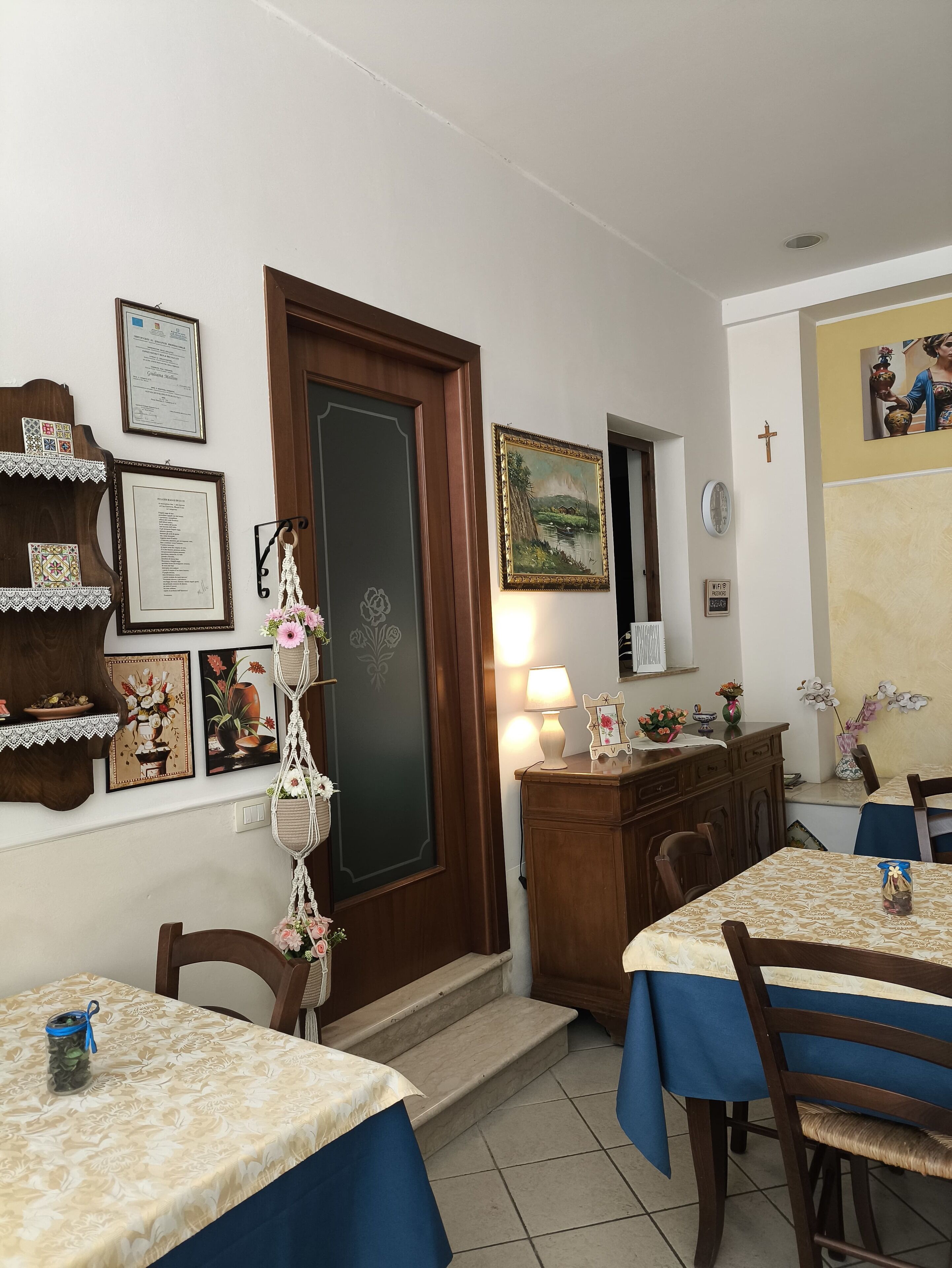 A dining area with wooden tables and chairs, a staircase leading to an upper level, and various framed certificates and artwork on the walls.