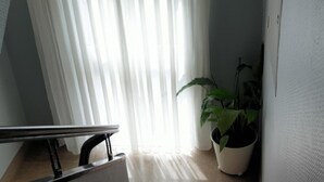 Una escalera con barandilla de metal, una planta en maceta y una ventana con cortinas translúcidas que dejan entrar luz natural.