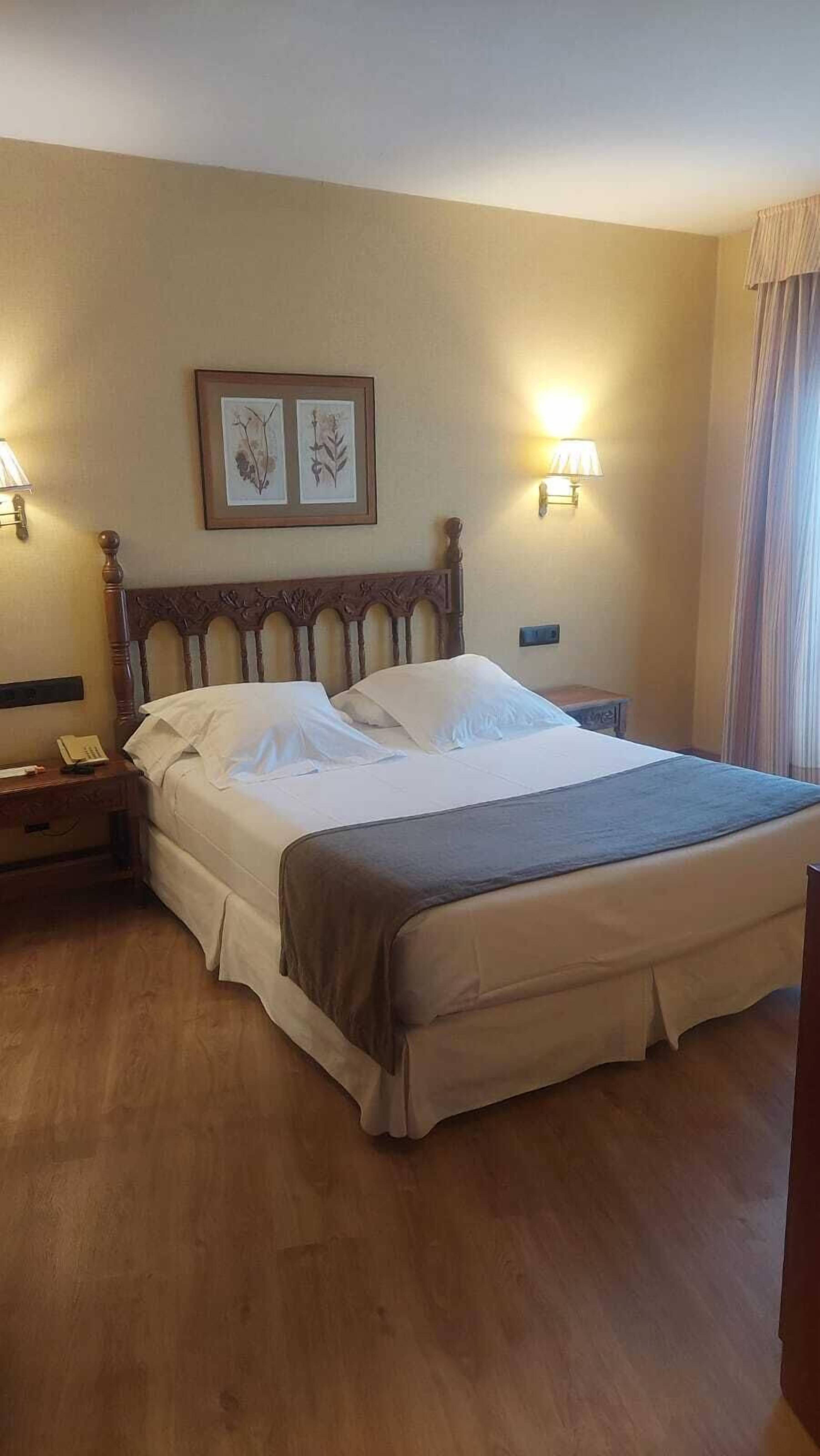 Una cama bien hecha con cabecera de madera, dos lámparas fijadas a la pared e impresión enmarcadas de motivos botánicos en la pared.
