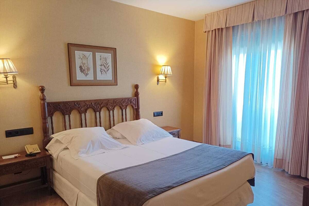 Une chambre d’hôtel avec un grand lit, deux lampes de chevet, une œuvre d’art florale encadrée au mur et une fenêtre avec des rideaux légers.