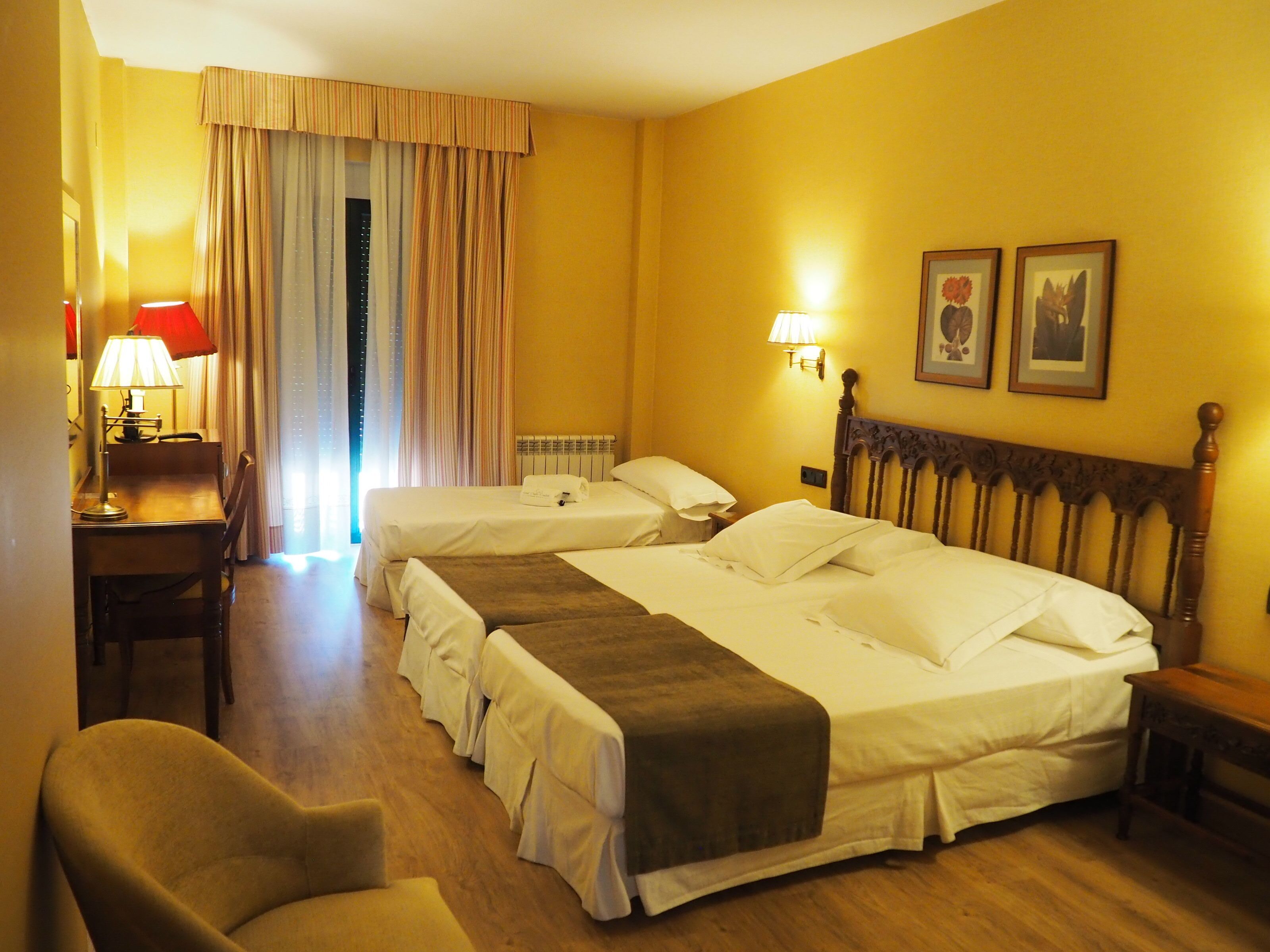 A hotel room with two beds, a desk with a lamp, a chair, and two wall-mounted lamps.