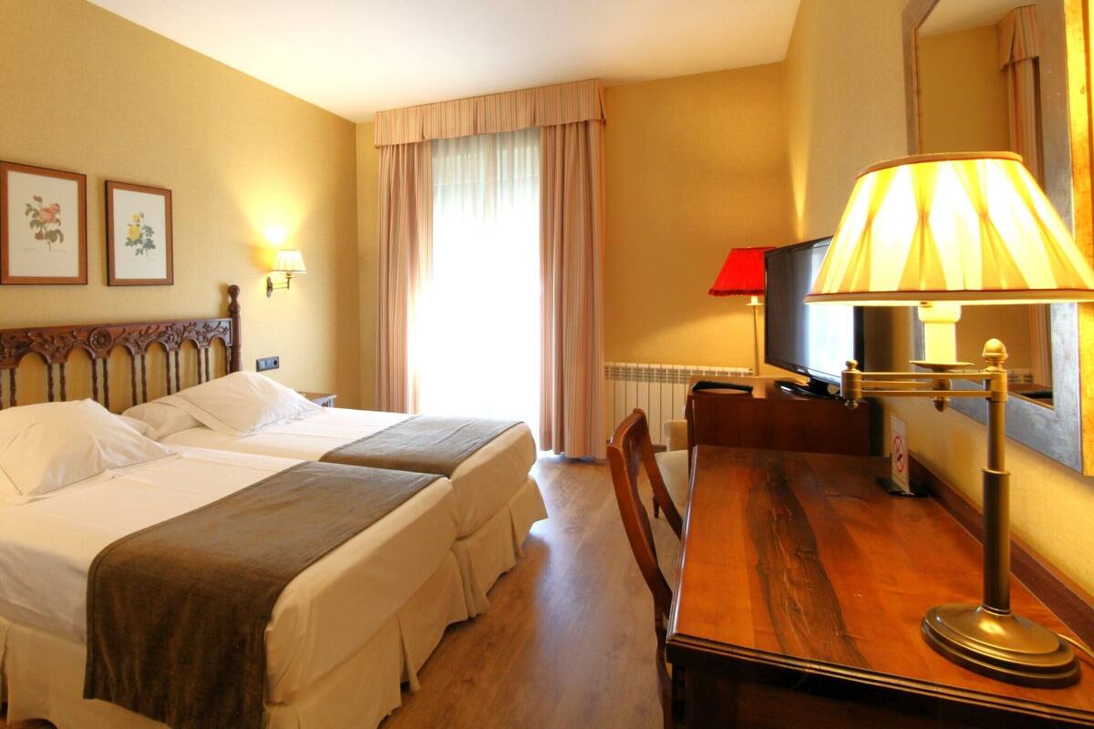 Une chambre d’hôtel avec un lit double, une table de chevet en bois, un bureau avec une télévision et deux tableaux représentant des motifs floraux, encadrés et accrochés au mur.