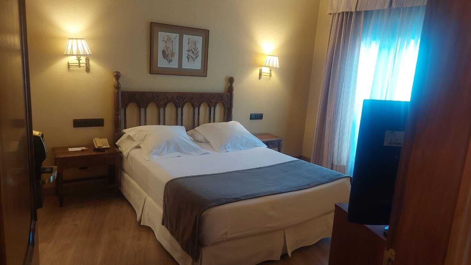 Quarto de hotel com cama de madeira, duas luminárias de cabeceira, uma moldura na parede e uma janela com cortinas.