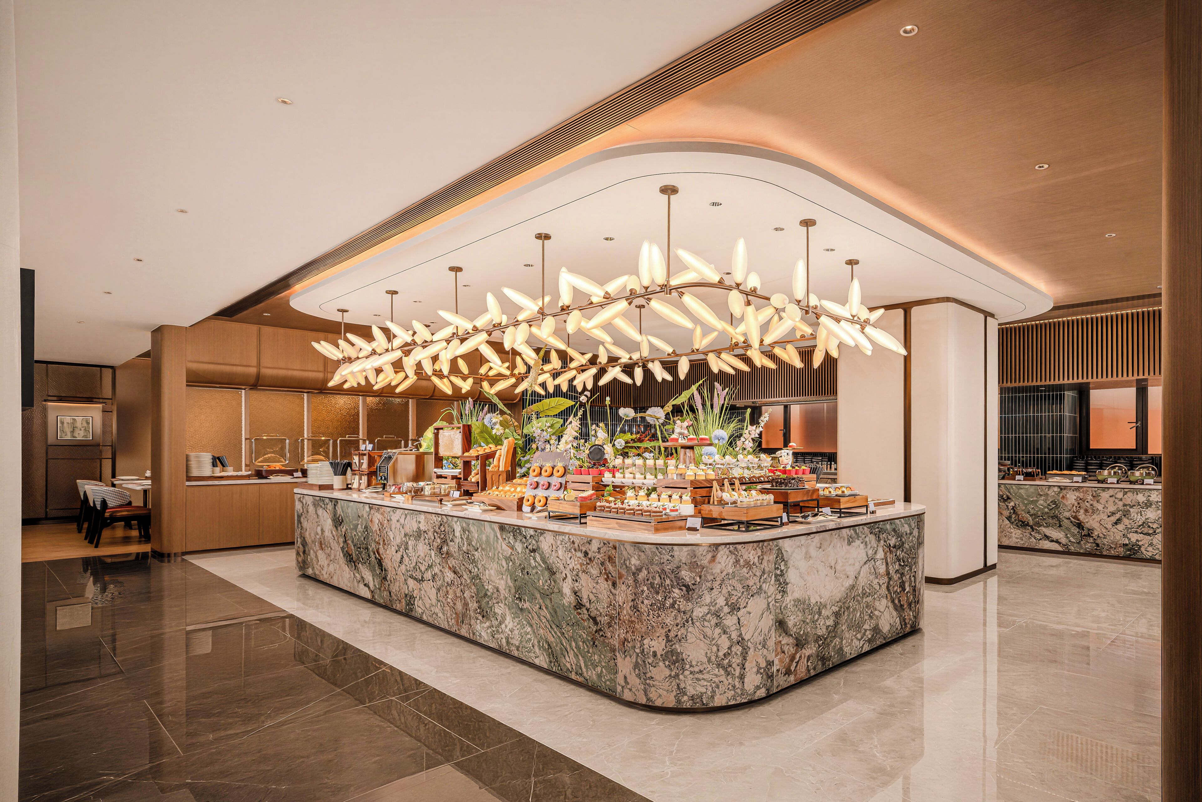 A modern hotel lobby with a marble reception desk, a large floral chandelier, and a buffet area.