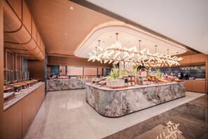A modern hotel lobby with a marble reception desk, buffet area, and a large decorative chandelier.