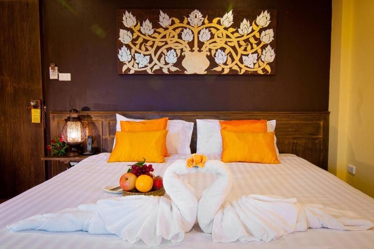 A hotel room with a large bed, two orange pillows, a decorative wall piece, a nightstand with a lamp, and a fruit arrangement.