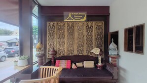 A room with a dark brown sofa, a wooden chair, and a decorative wall panel. A sign above the sofa reads "CM Night Bazaar Boutique Hotel."