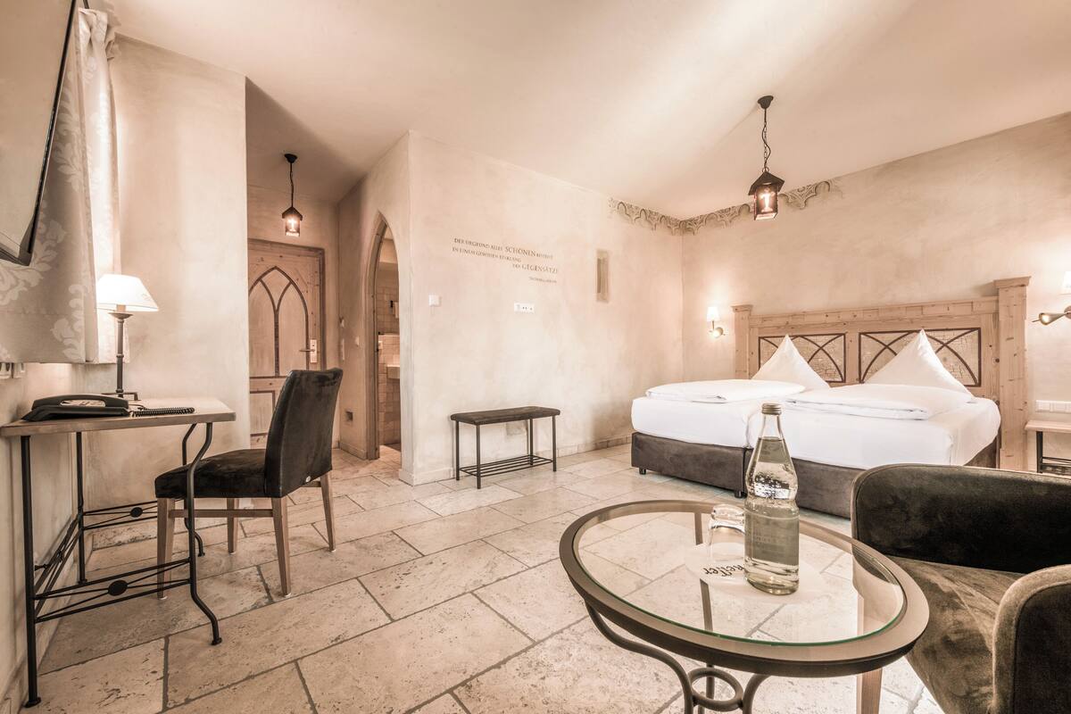 A hotel room with a bed, a desk with a chair, a small table, and a lamp.