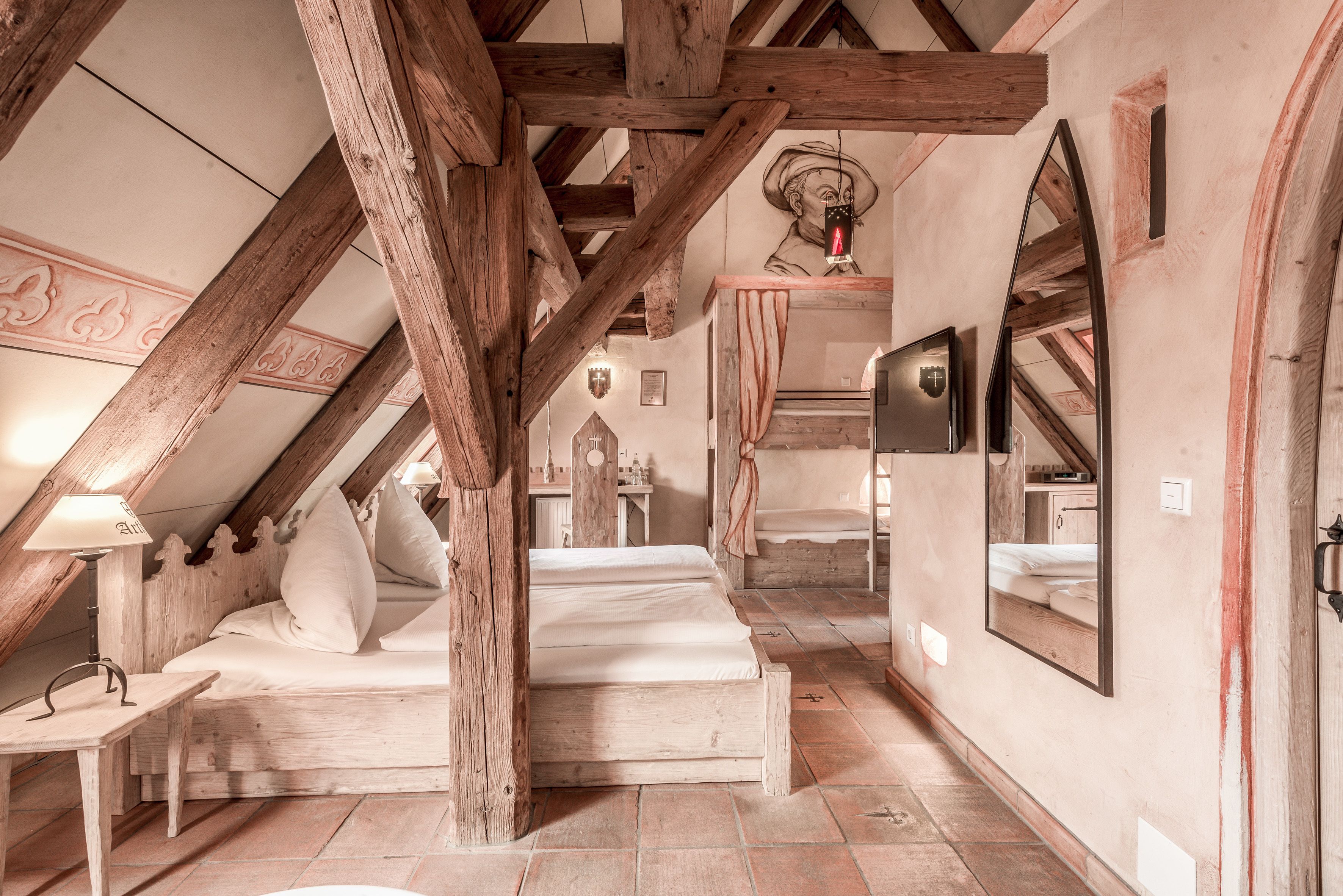 A rustic attic bedroom with exposed wooden beams, a bed with white linens, a small table with a lamp, and a mirror on the wall.