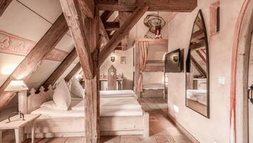 A rustic attic bedroom with exposed wooden beams, a bed with white linens, a small table with a lamp, and a mirror on the wall.