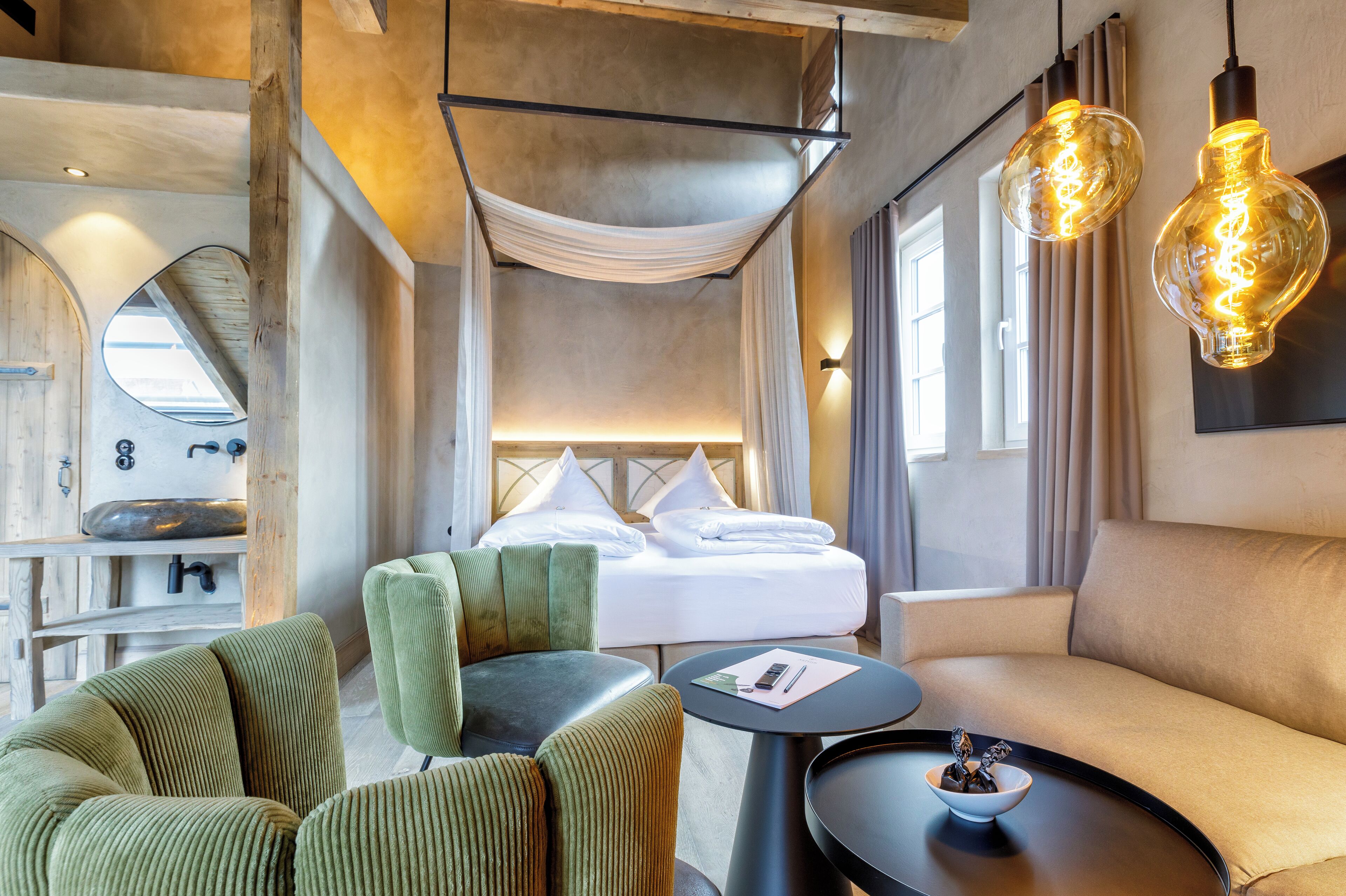 Habitació d'hotel moderna amb un llit, cadires tapissades de verd, una taula rodona i un llum penjant.