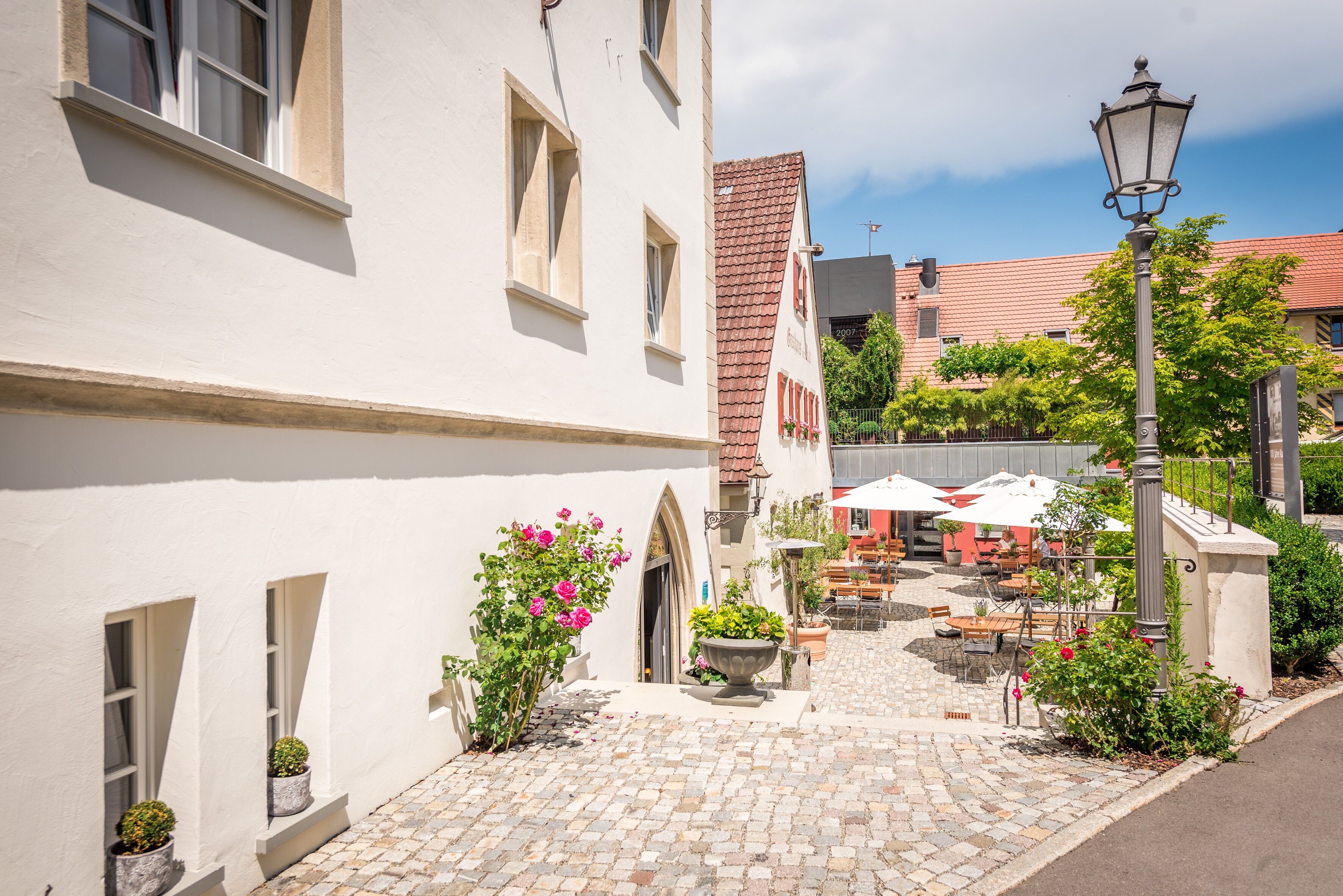 Une cour pavée, avec un bâtiment blanc, une construction à toit rouge, des sièges en plein air et un lampadaire.
