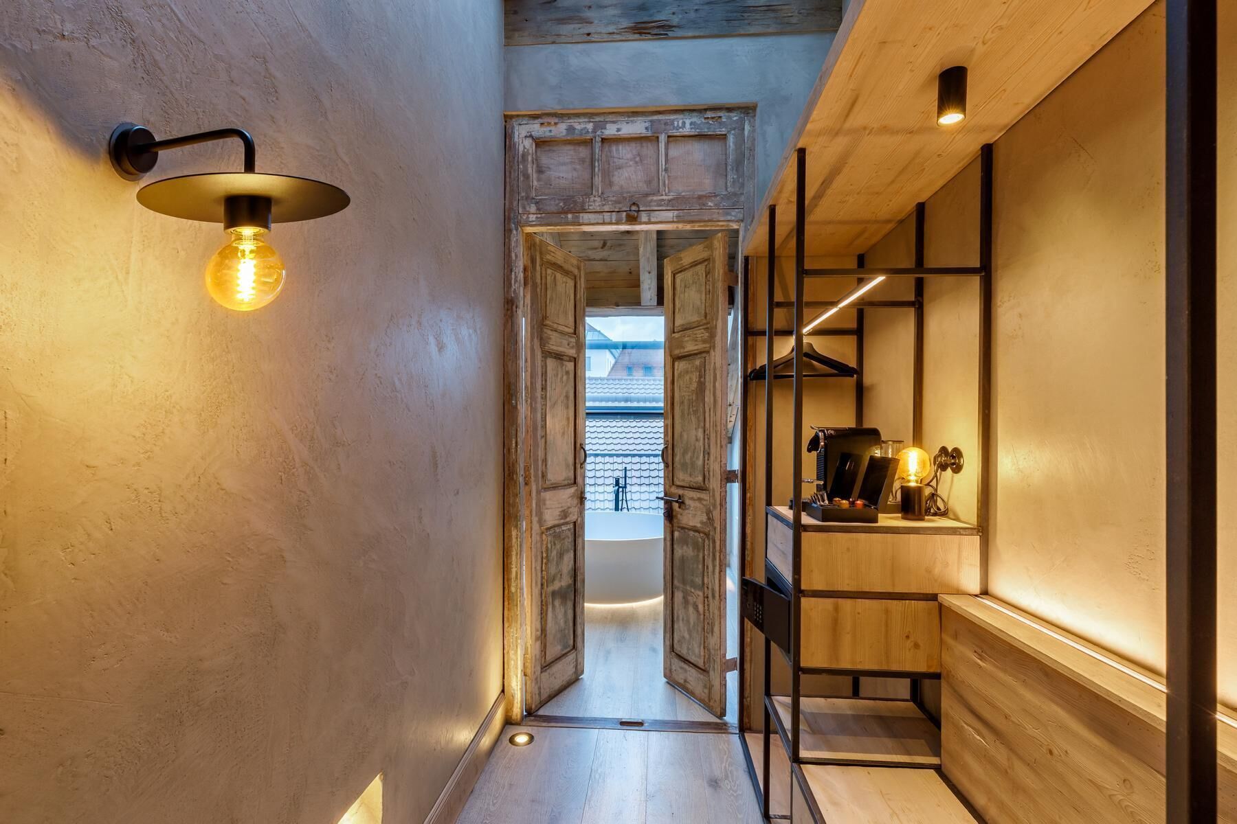 Een smalle gang met een houten deur die uitkomt op een balkon, een wandlamp en een kleine tafel met een lamp en een kop.