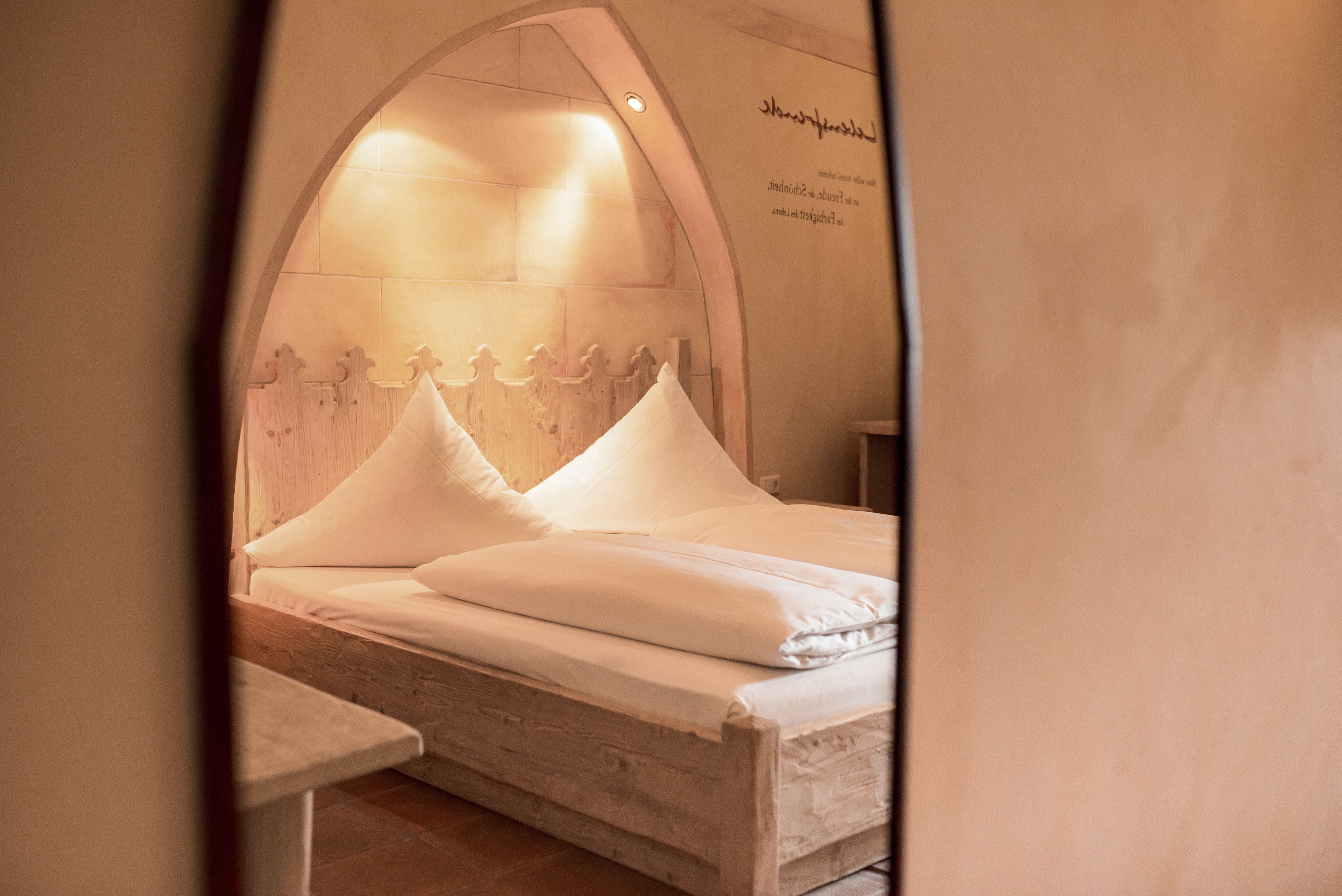 Ein Bett mit Holz-Kopfstütze und weißer Bettwäsche in einem Raum mit Fliesenboden und einer gewölbten Wand.