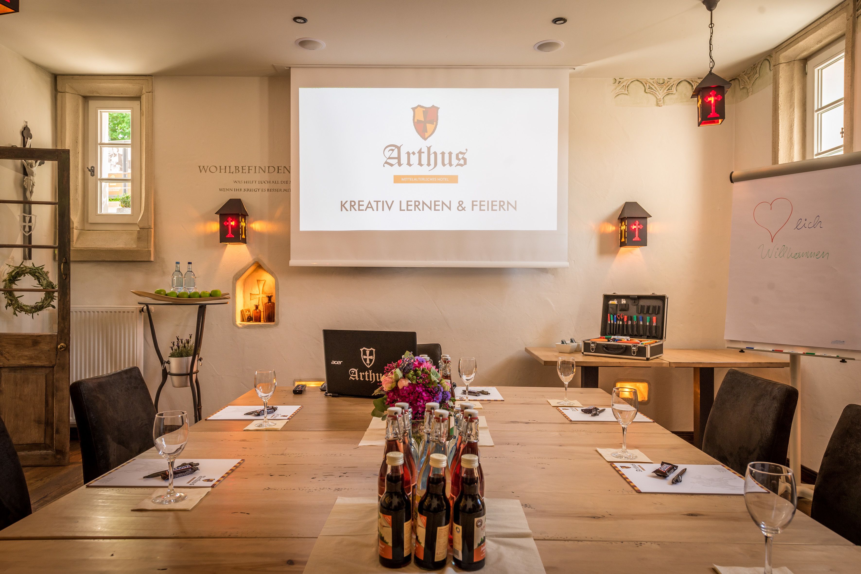 Uma sala de conferência com uma tela de projetor exibindo "Arthus Kreativ Lernen & Feiern", uma mesa de madeira disposta com garrafas de vinho e copos, e uma lousa com a palavra "Wohlbefinden" escrita.