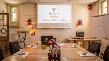 Uma sala de conferência com uma tela de projetor exibindo "Arthus Kreativ Lernen & Feiern", uma mesa de madeira disposta com garrafas de vinho e copos, e uma lousa com a palavra "Wohlbefinden" escrita.