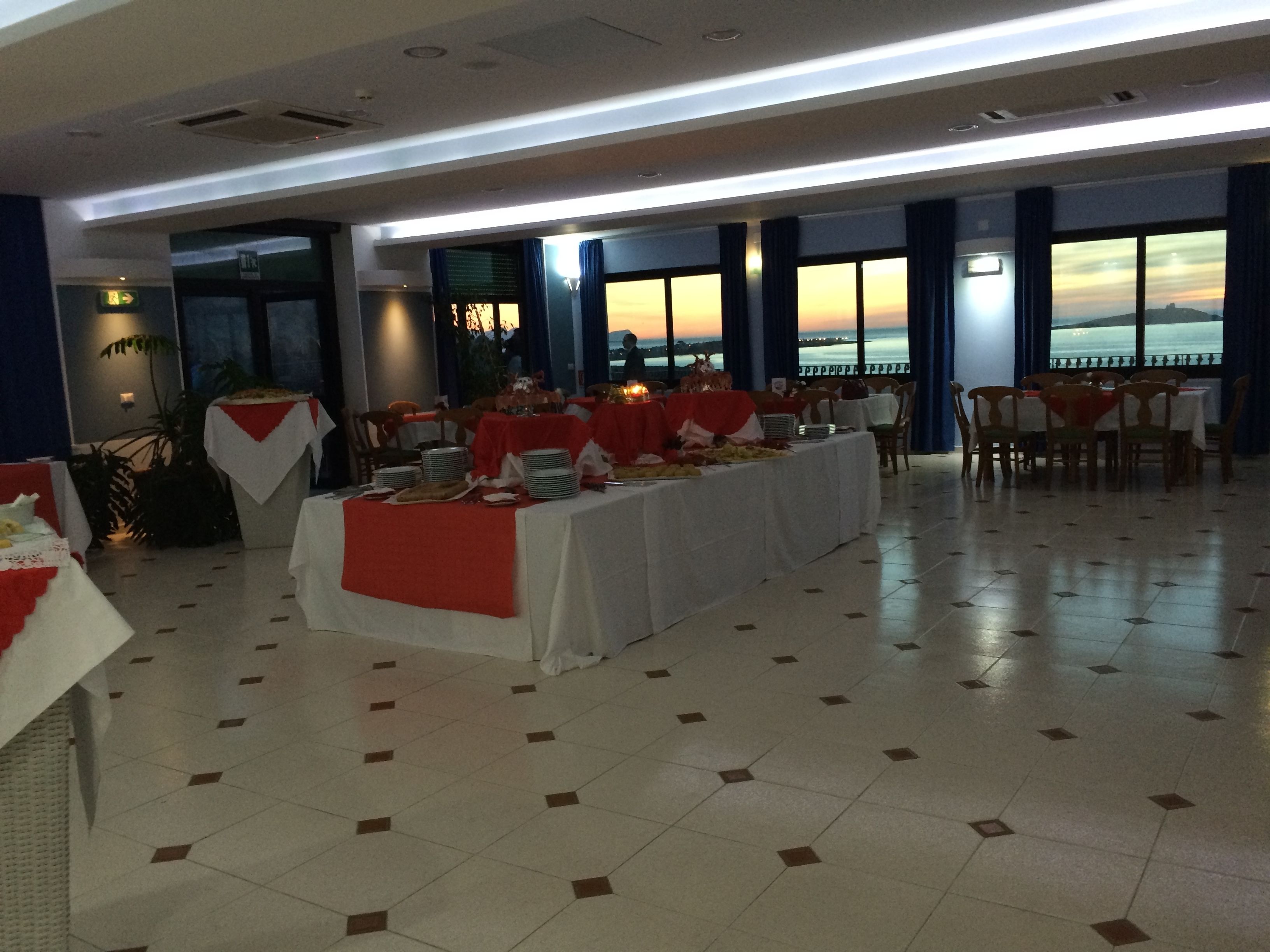 A banquet hall with a long table set for a meal, large windows with a sunset view, and a tiled floor.