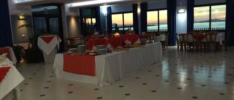 A banquet hall with a long table set for a meal, large windows with a sunset view, and a tiled floor.