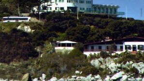 Um edifício à beira-mar com vários andares, rodeado por terreno rochoso e vegetação.