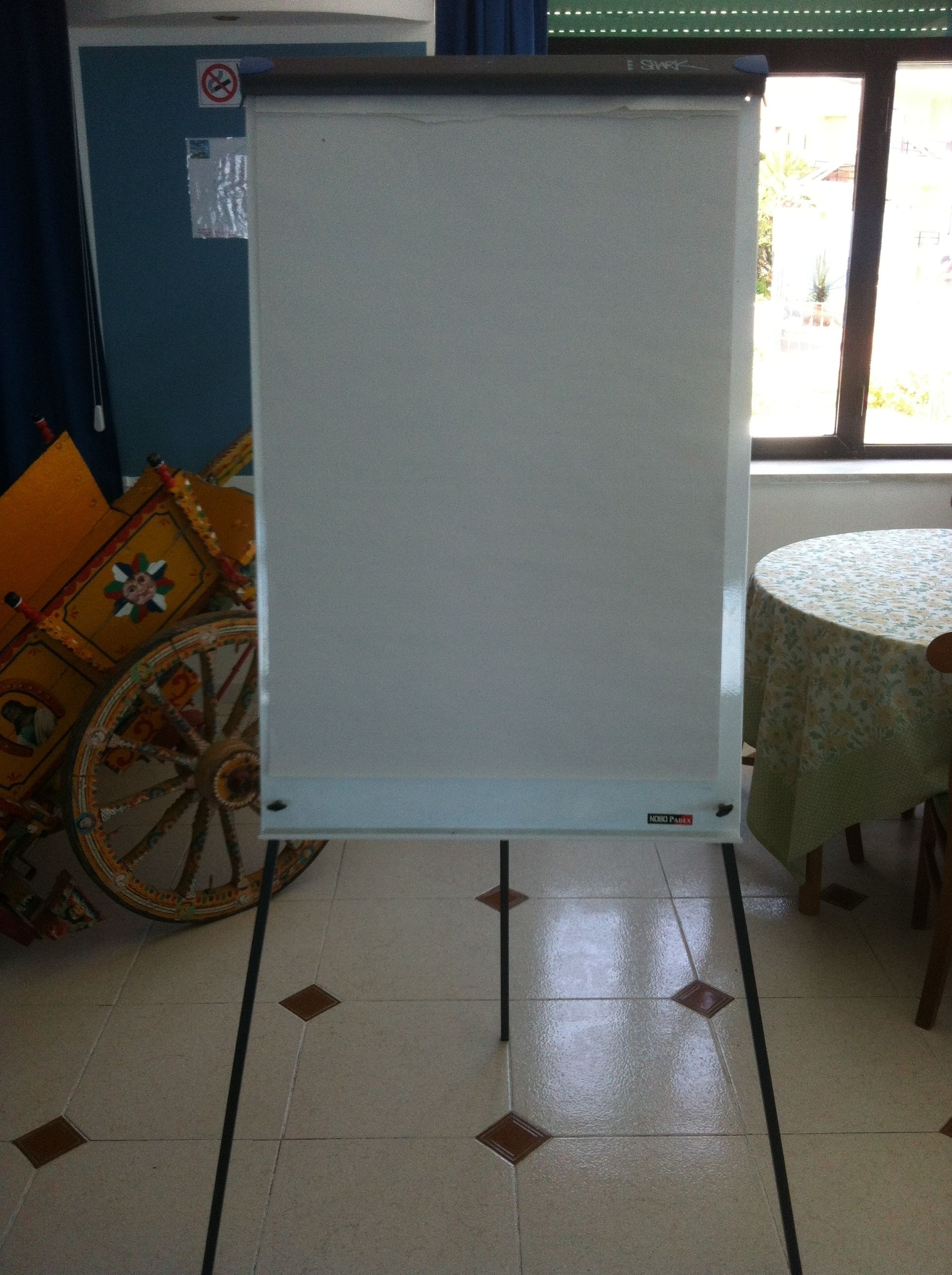 A room with a whiteboard stand, a table with a floral tablecloth, and a decorative wheel.