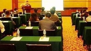 A conference room with rows of tables, a projector screen, and a speaker at the front.
